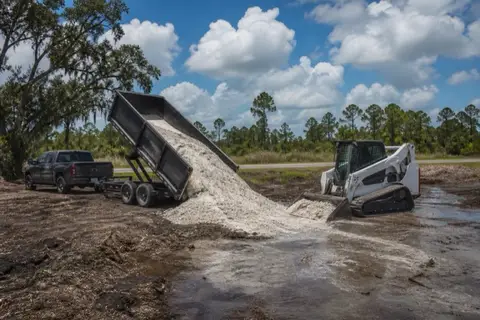 Dump trailer tipping pale sand fill onto the low corner of a cleared Palm Coast lot as a compact loader pushes it out toward the swale line