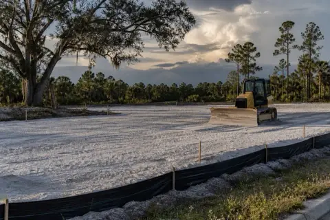 Small dozer spreading pale sand across a cleared building pad with grade stakes and string lines set and a silt fence along the street swale