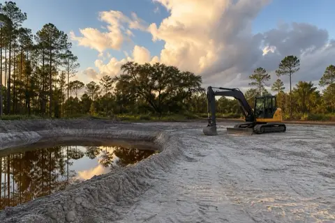 Freshly excavated stormwater pond holding tea-colored water beside a graded building pad with a compact excavator parked at golden hour